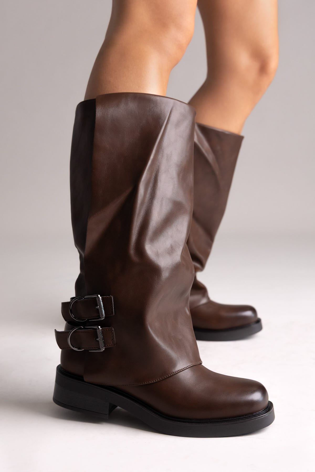 Model legs in brown leather knee-high boots with buckle straps and chunky sole, angled view on a light grey background