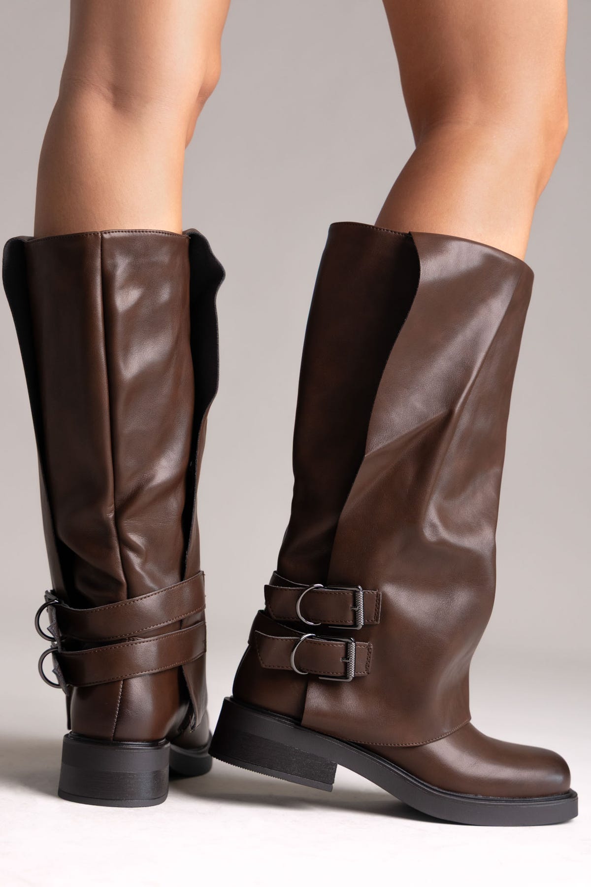 Model legs in brown leather knee-high boots with buckle straps and chunky sole, angled view on a light grey background