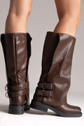Model legs in brown leather knee-high boots with buckle straps and chunky sole, angled view on a light grey background