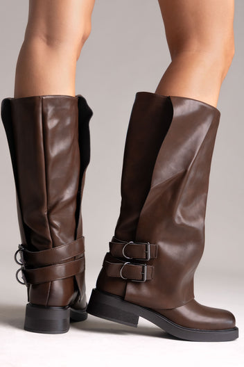 Model legs in brown leather knee-high boots with buckle straps and chunky sole, angled view on a light grey background