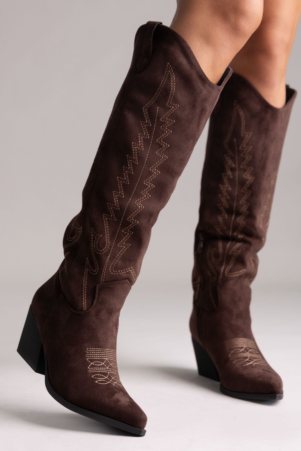 Dark brown suede knee-high western boots with stitched shaft detail and pointed toe, side view on a model