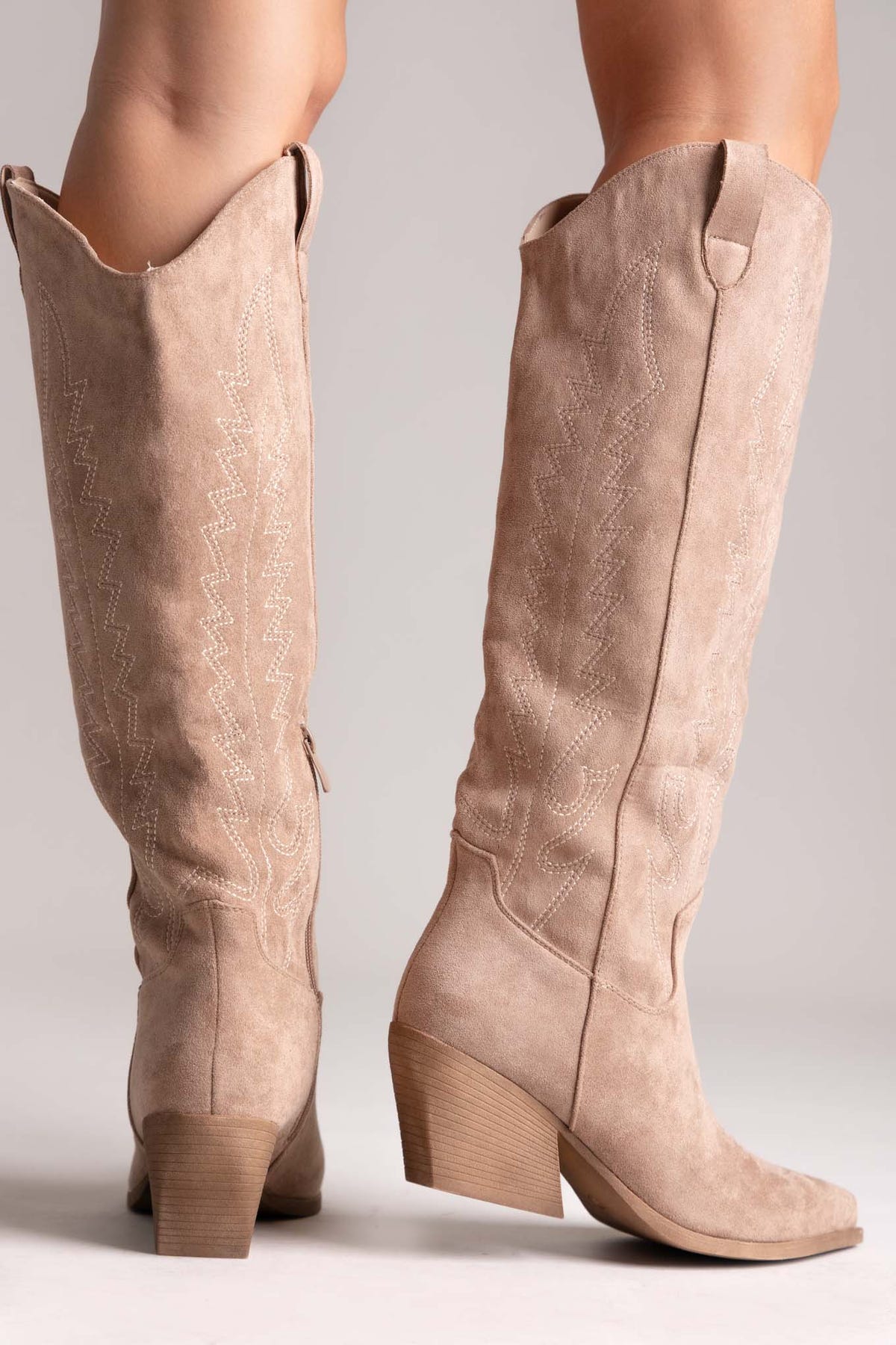 Taupe suede knee-high western boots with stitched shaft detail and pointed toe, rear view on a model