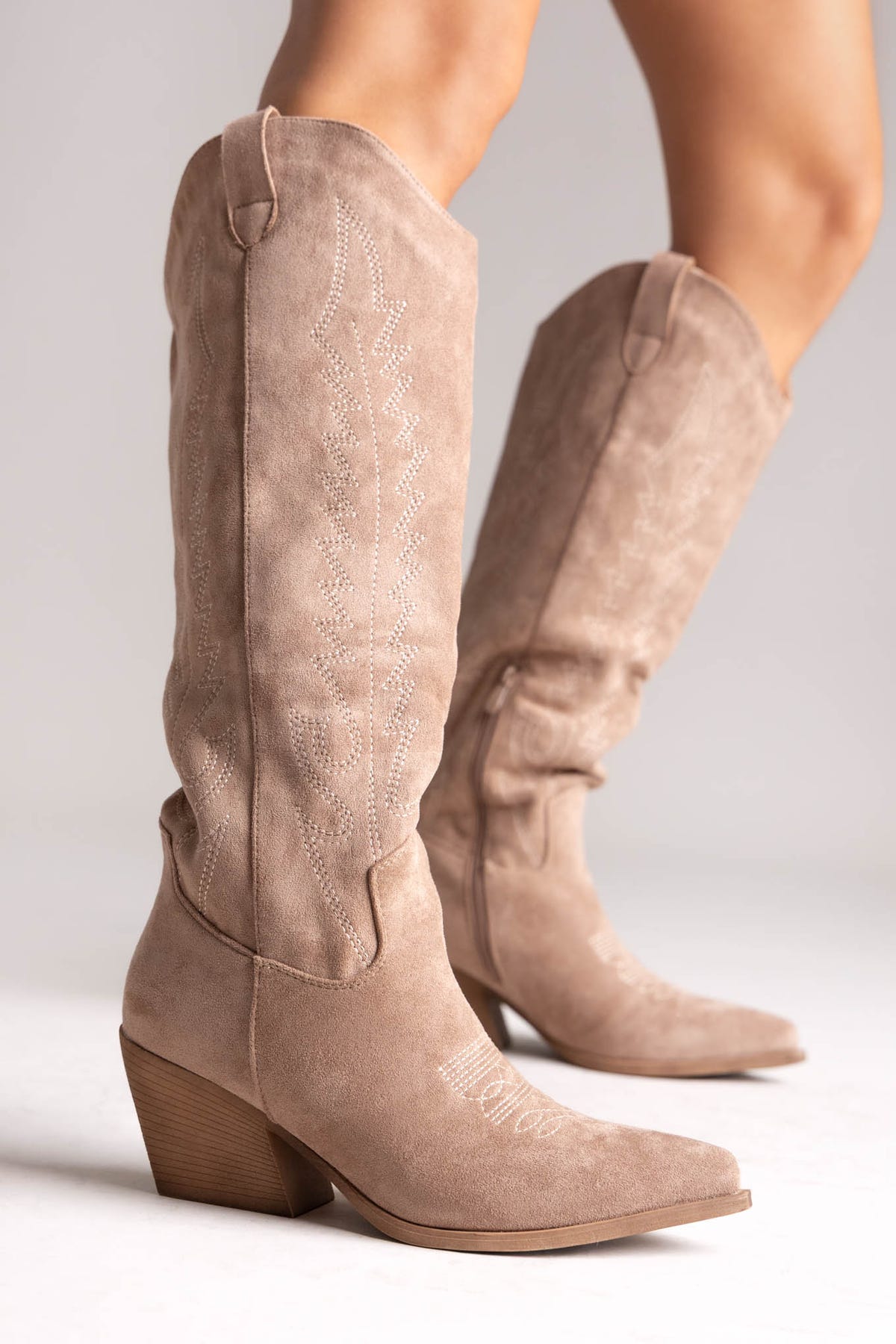 Taupe suede knee-high western boots with stitched shaft detail and pointed toe, angled view on a model