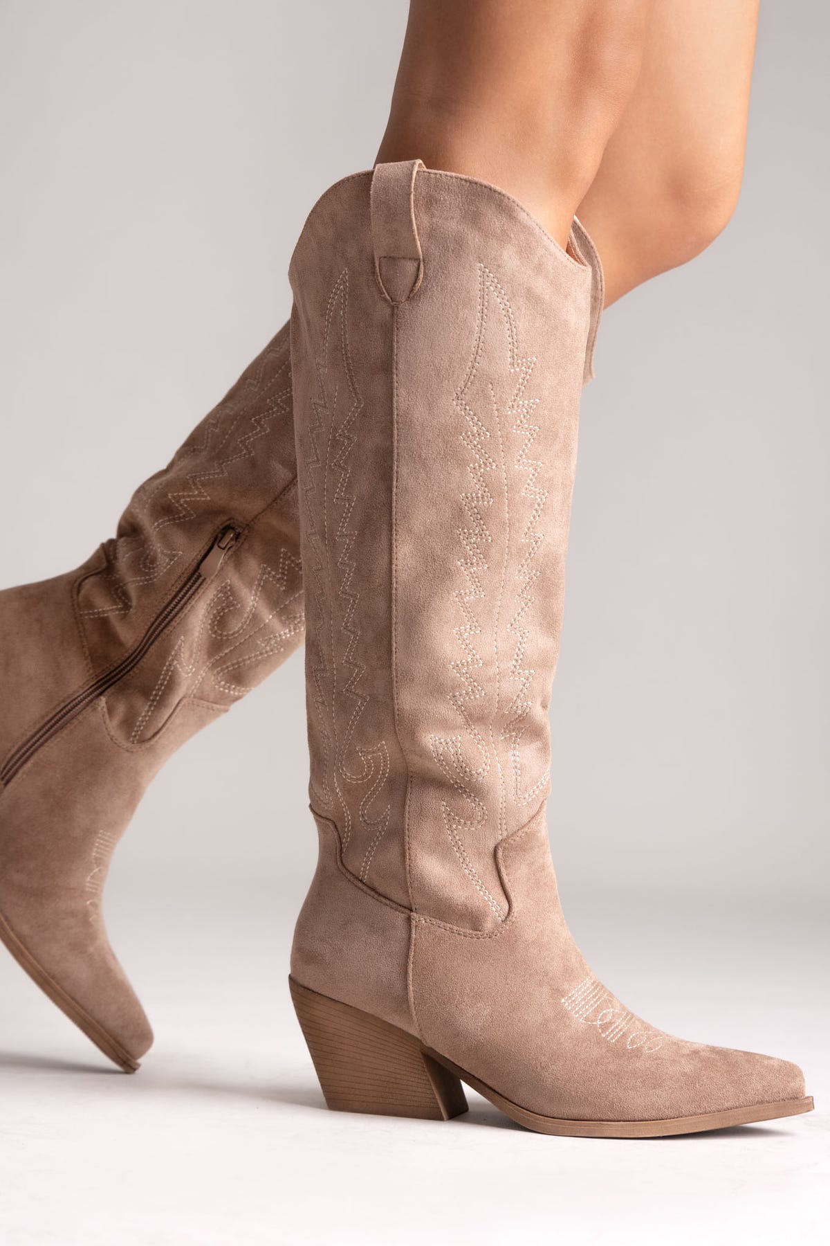 Taupe suede knee-high western boots with stitched shaft, side zip and pointed toe, side view on a model