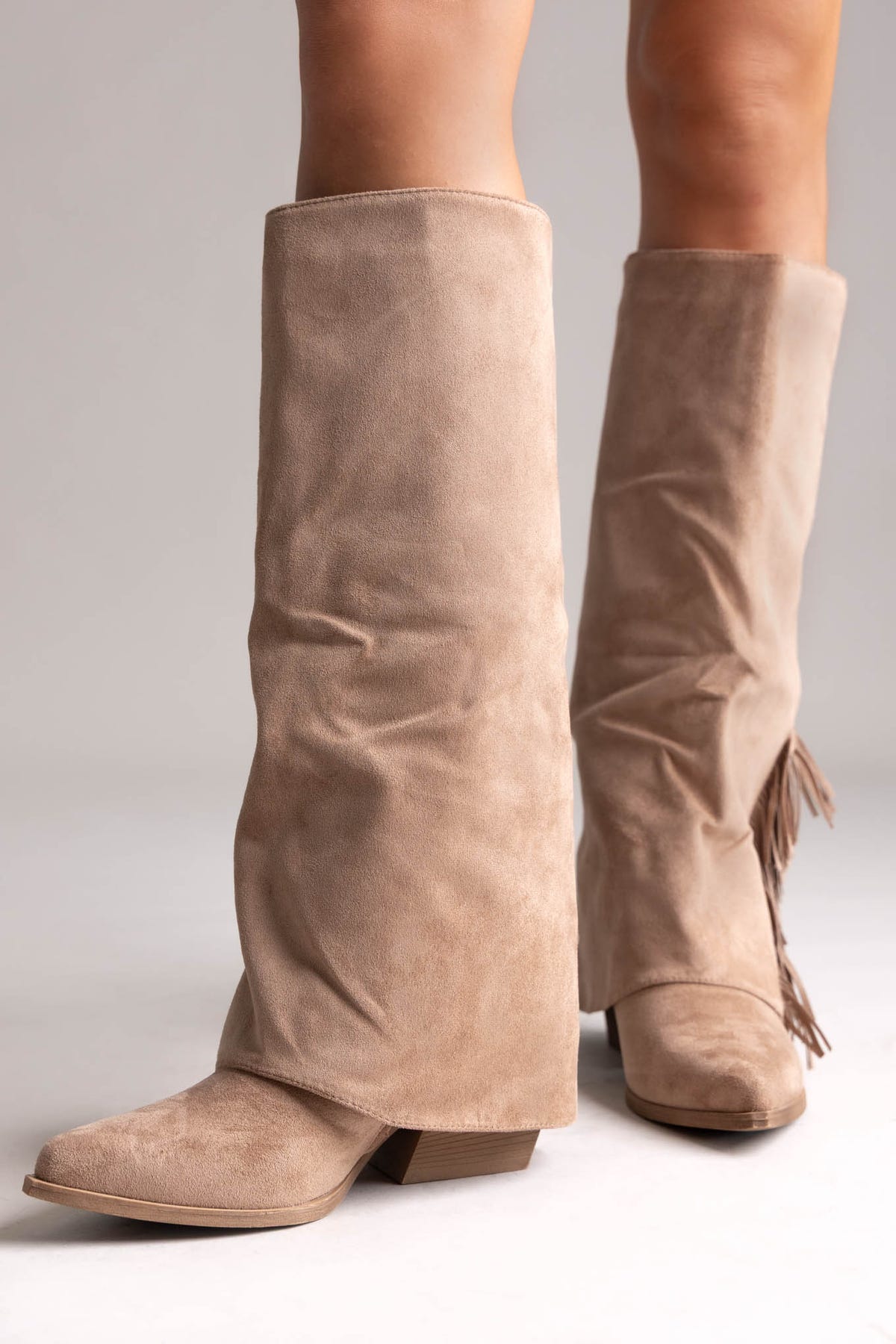 Taupe suede fold-over knee-high boots with fringe trim along the side and block heel, side view on a model
