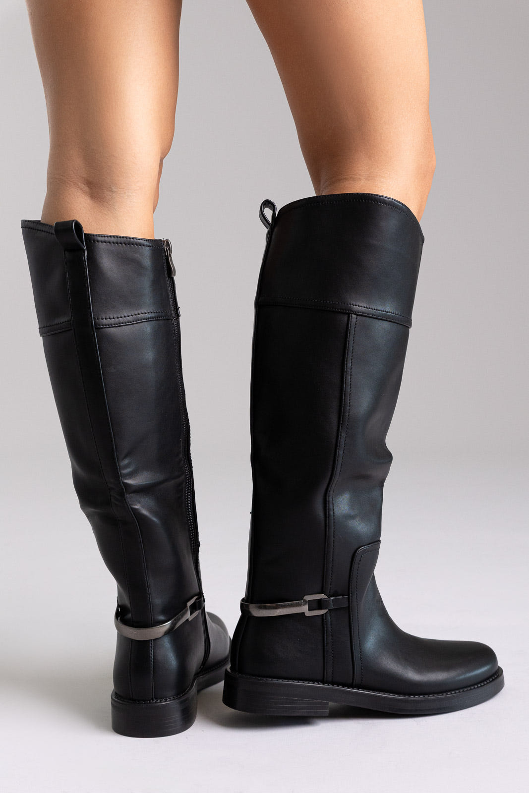 Black faux leather knee-high riding boots with silver buckle detail and flat sole, angled view on a model