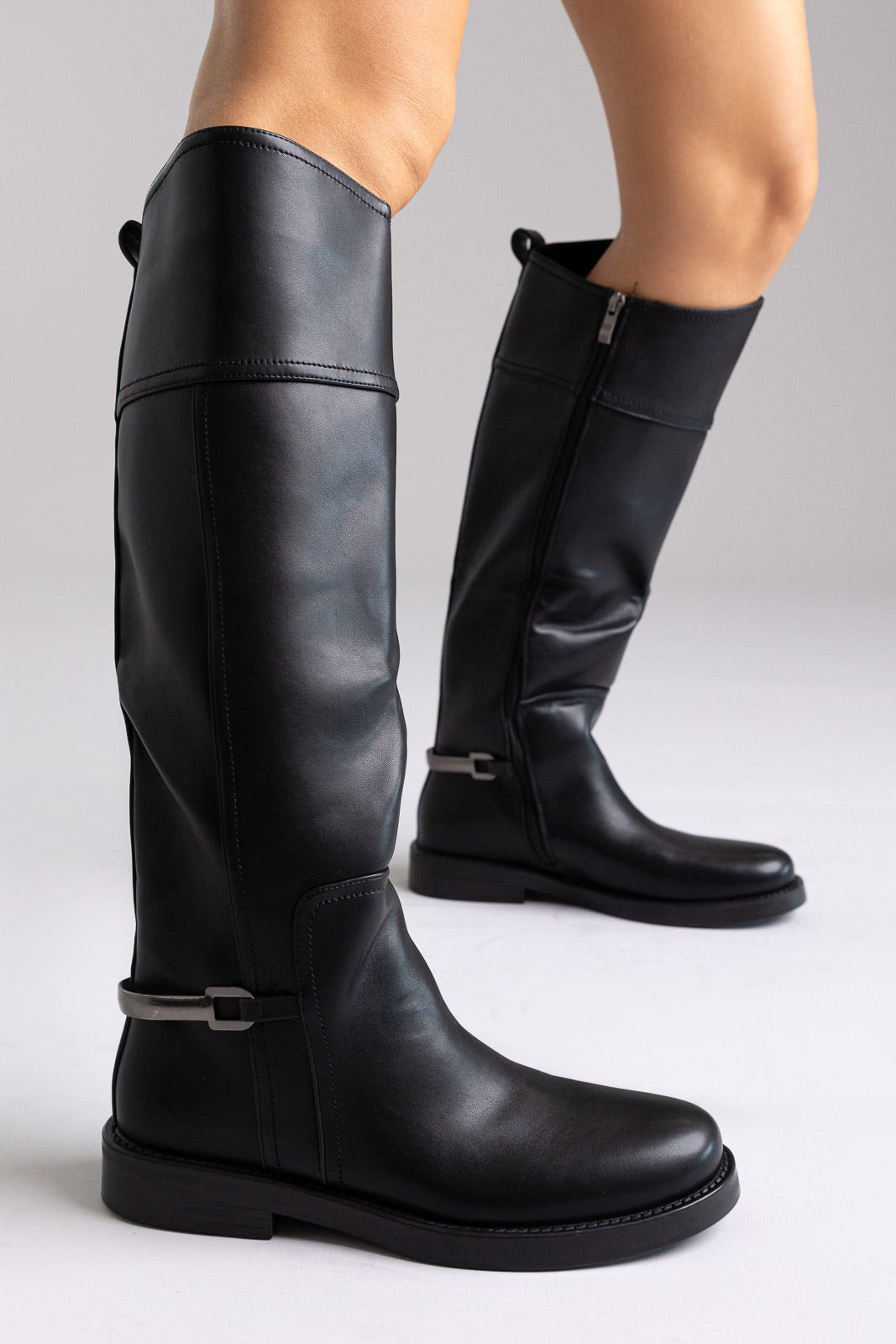 Black faux leather knee-high riding boots with buckle detail and flat sole, angled view on a model