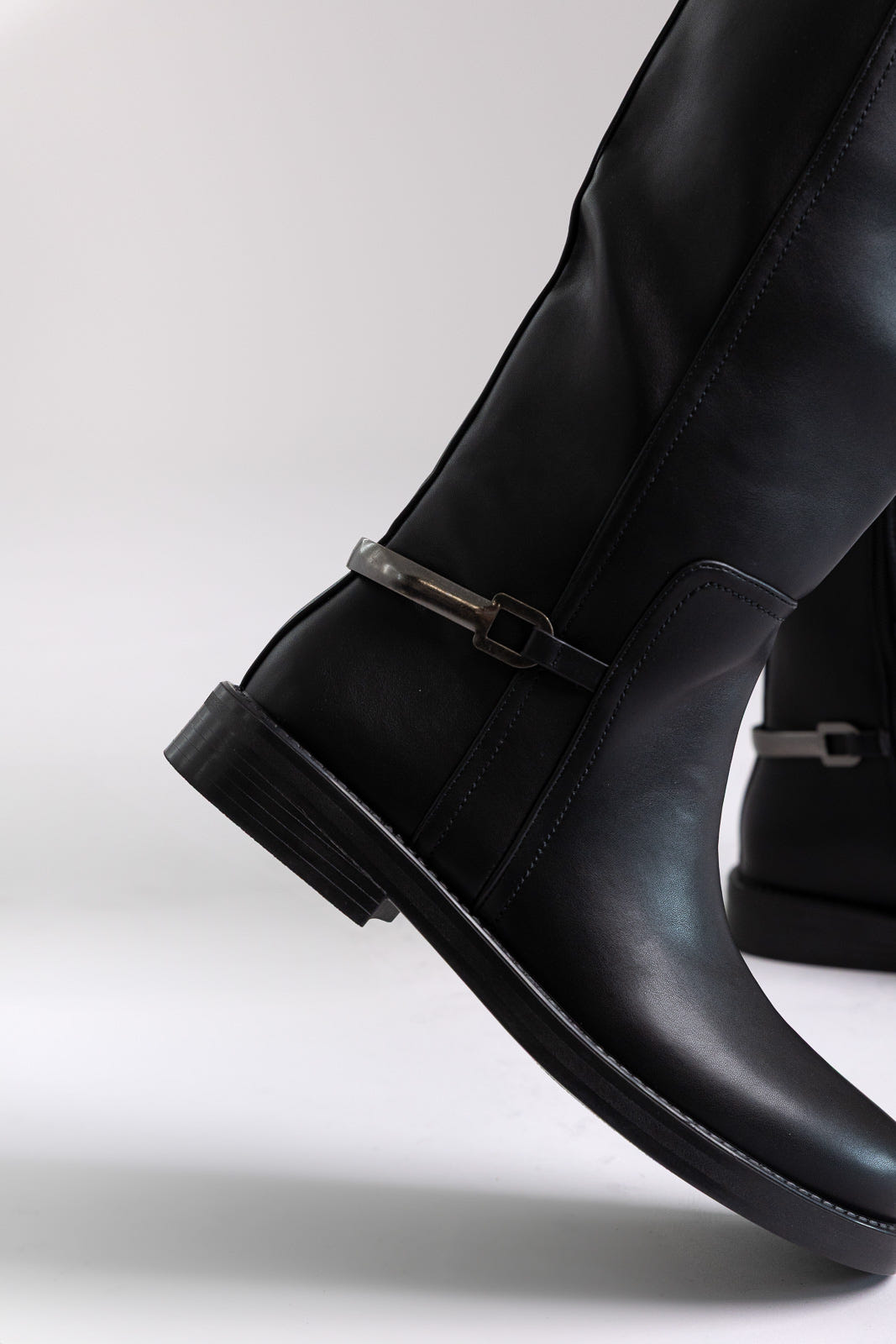 Black faux leather knee-high riding boots with buckle detail and flat sole, close-up on a model