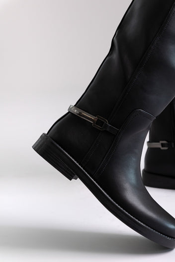 Black faux leather knee-high riding boots with buckle detail and flat sole, close-up on a model