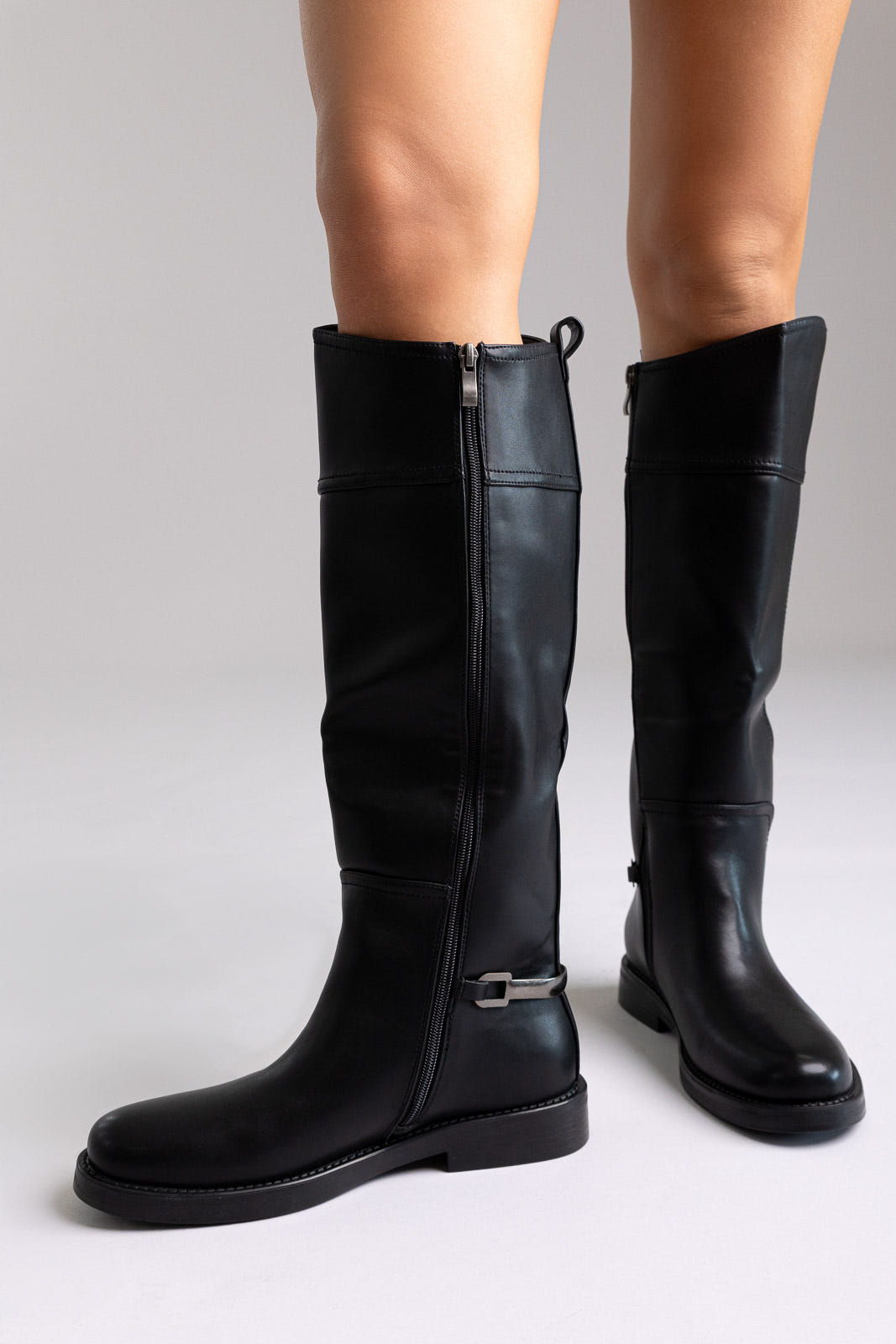 Black faux leather knee-high riding boots with buckle strap and flat sole, angled view on a model