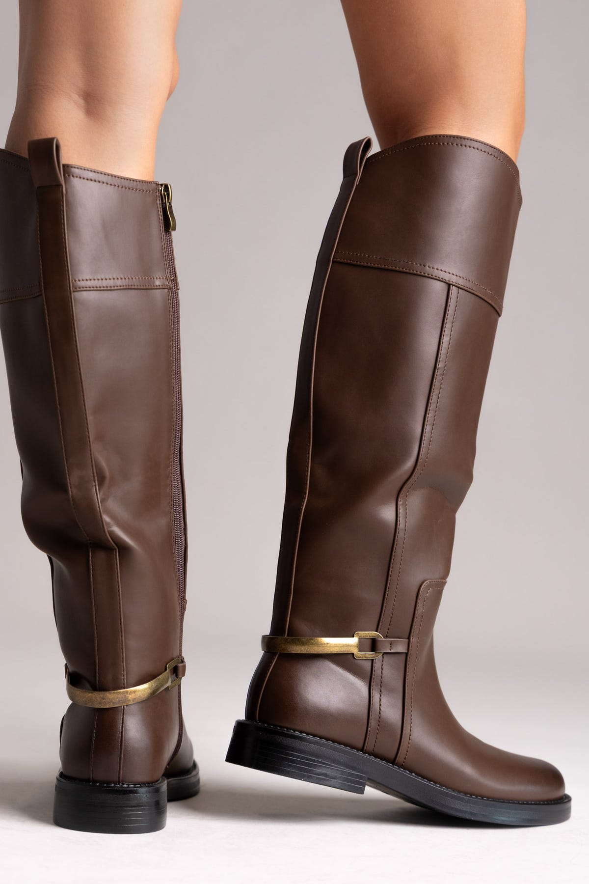 Brown leather knee-high riding boots with gold buckle strap and flat sole, rear view on a model