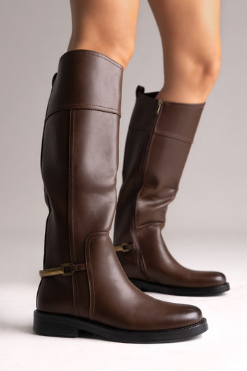 Brown leather knee-high riding boots with gold buckle strap and flat sole, side view on a model