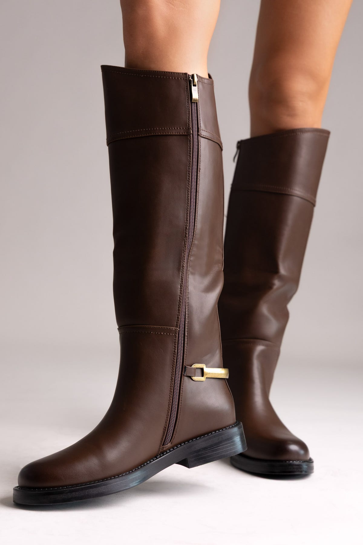 Brown leather knee-high riding boots with flat sole and side zip, side view on a model