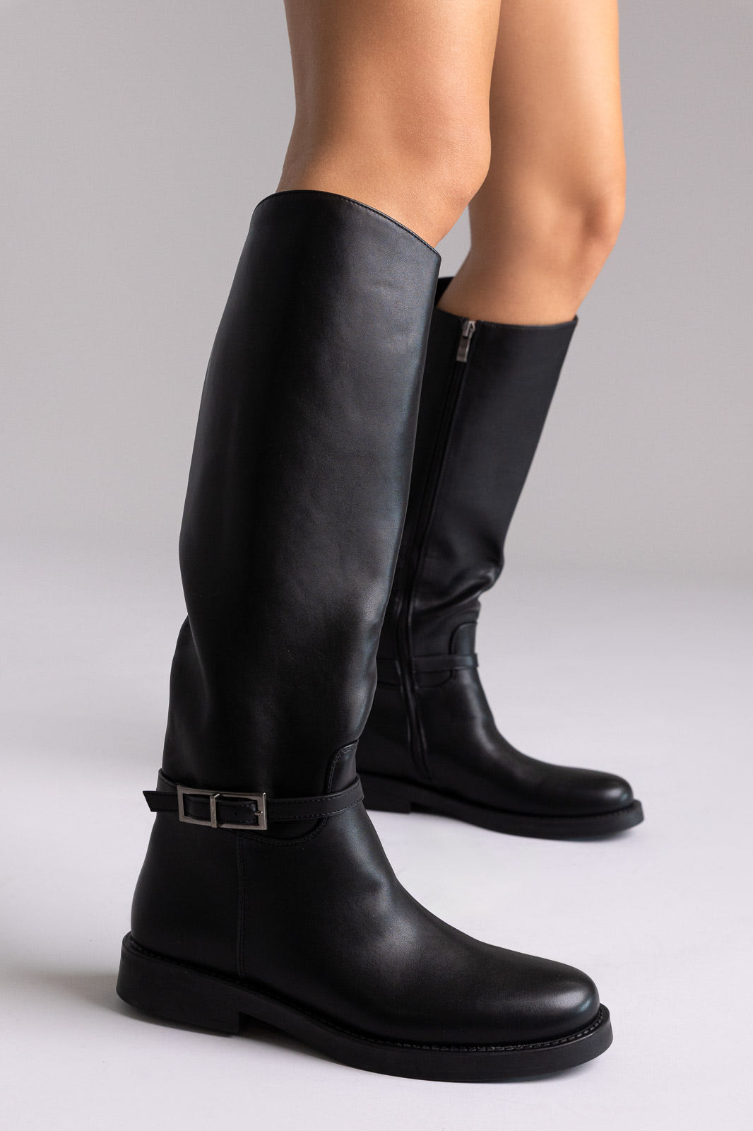 Black faux leather knee-high riding boots with buckle strap and flat sole, angled view on a model