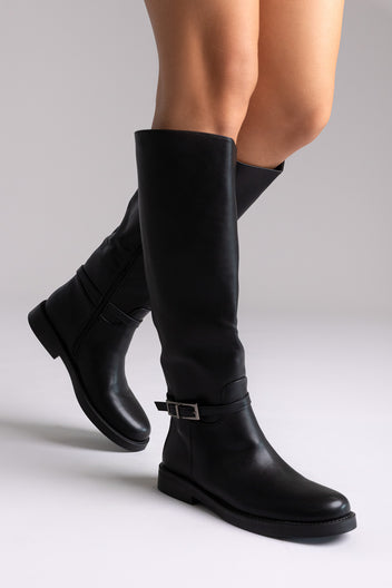 Black faux leather knee-high riding boots with buckle strap and flat sole, angled view on a model
