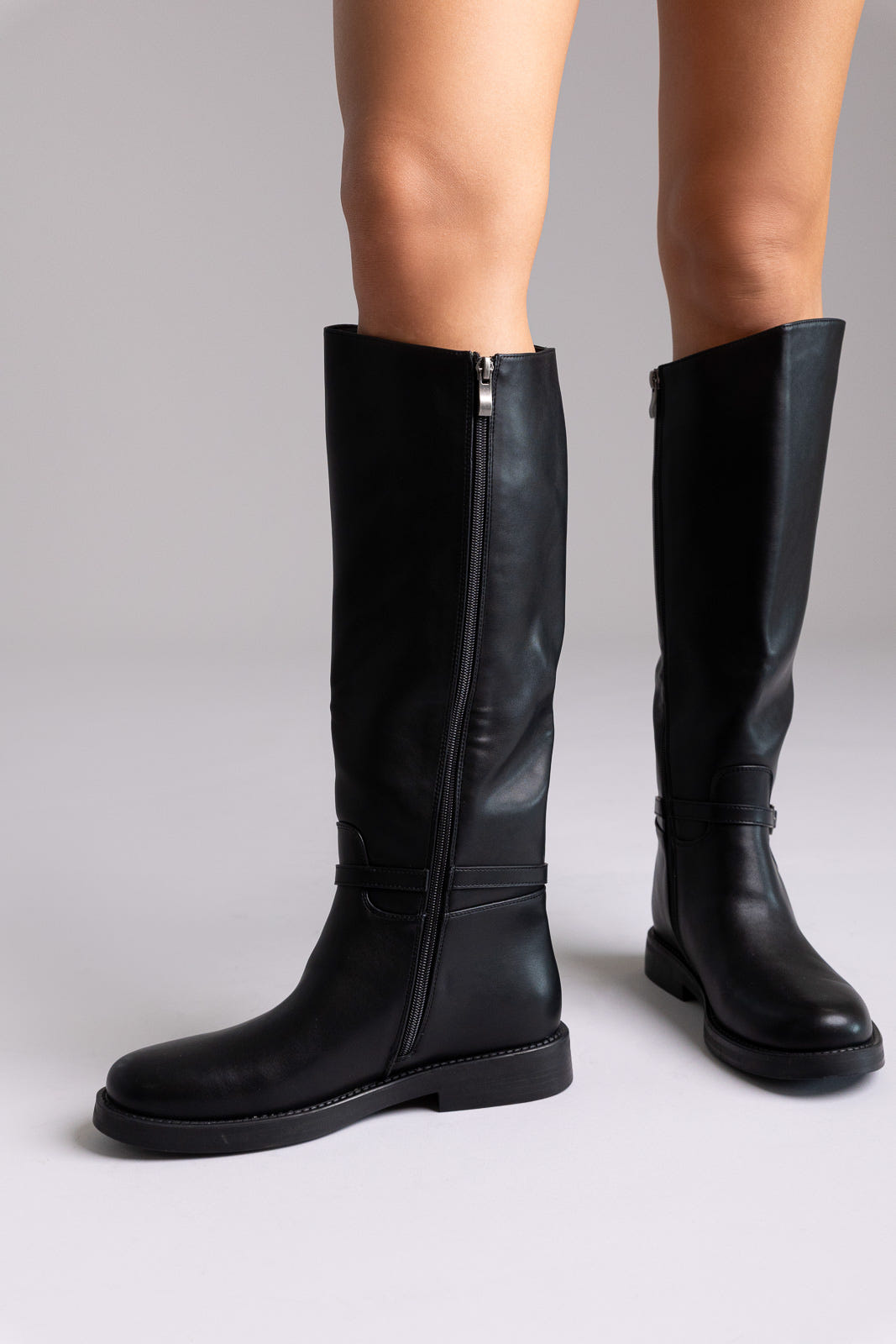 Black faux leather knee-high riding boots with side zip and flat sole, angled view on a model