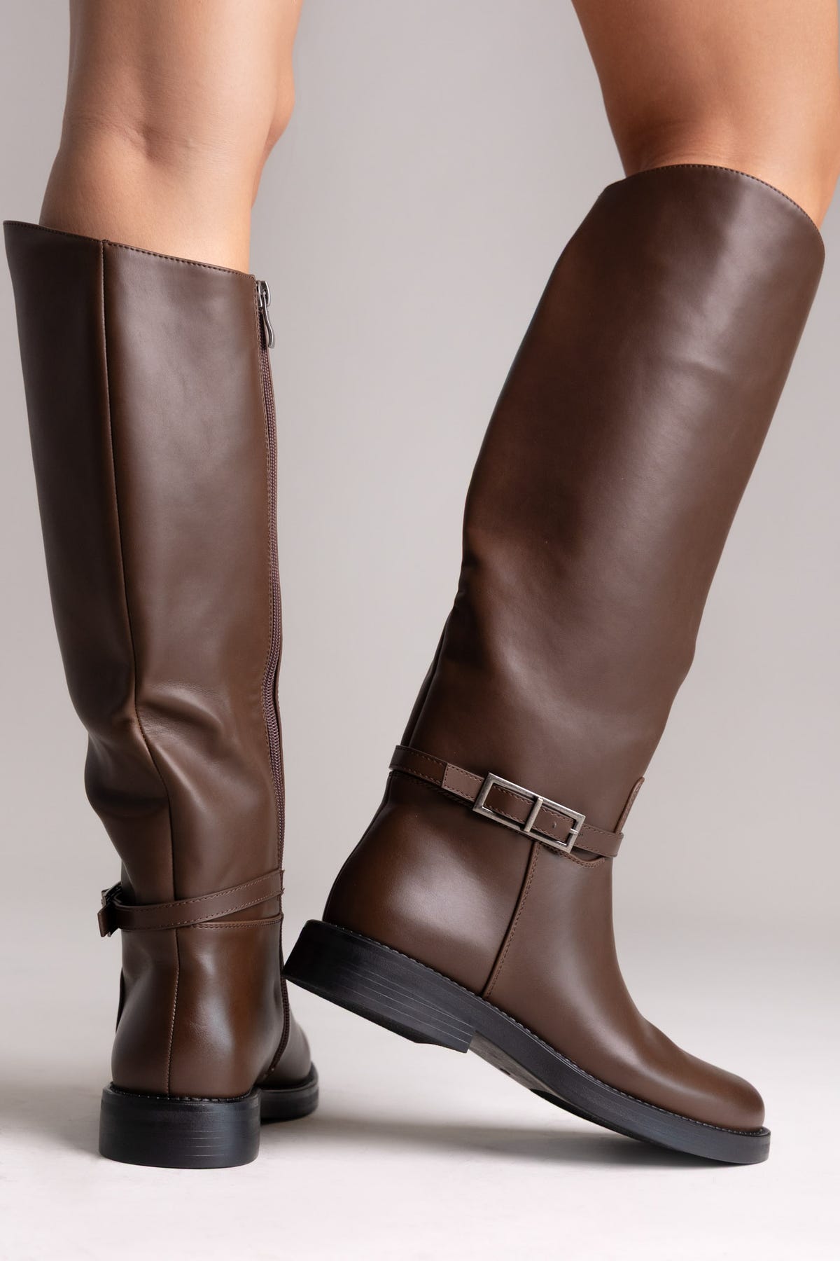 Brown leather knee-high riding boots with buckle strap and flat sole, rear angled view on a model