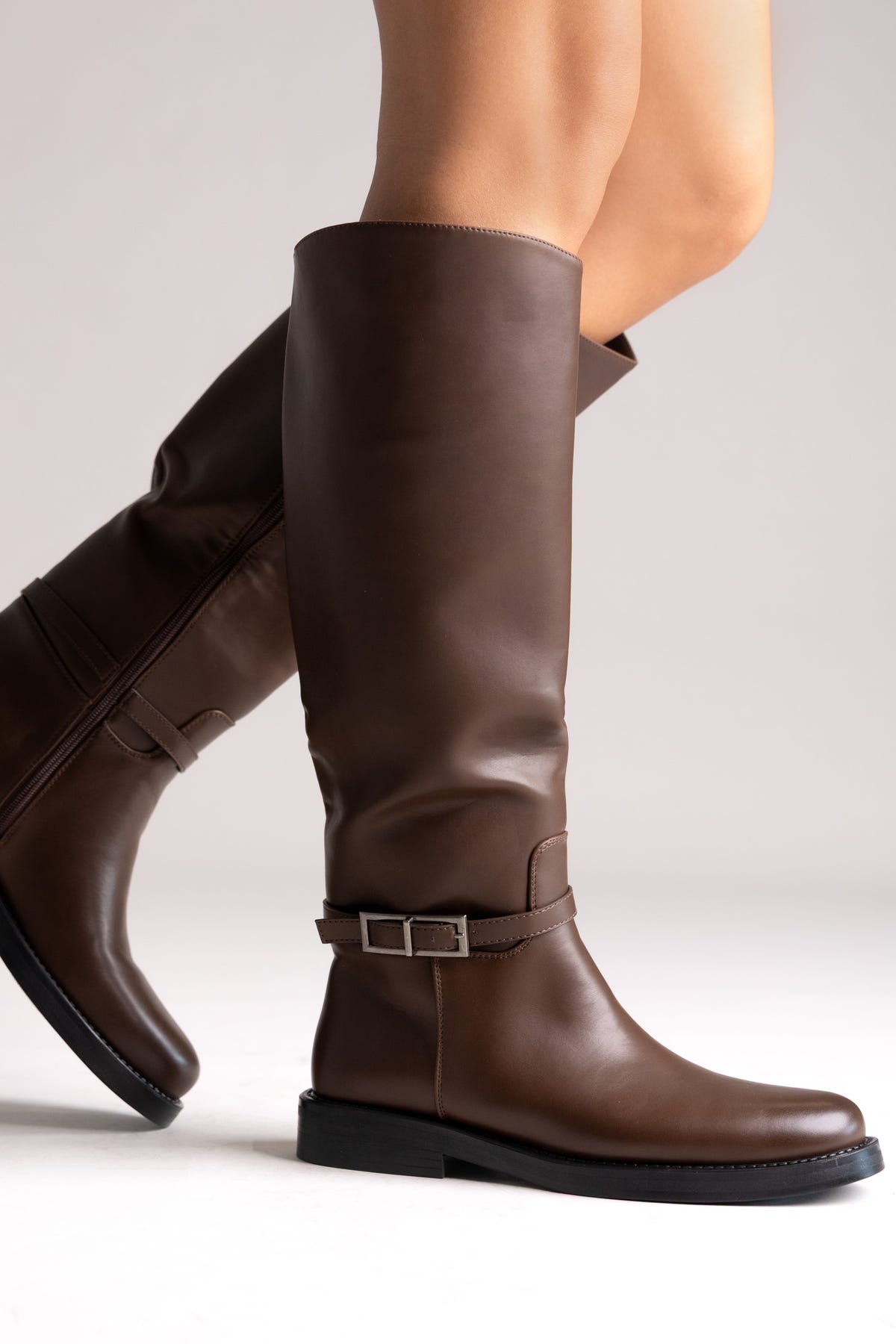 Brown leather knee-high riding boots with buckle strap and flat sole, angled view on a model