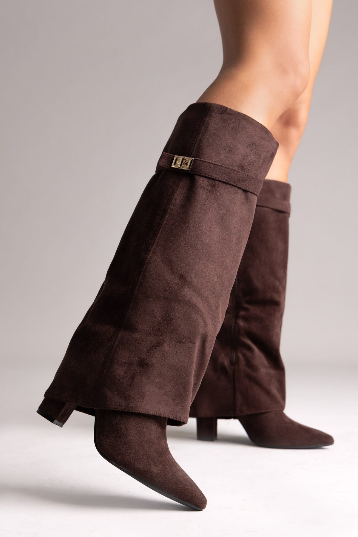 Dark brown faux suede fold-over knee-high boots with gold buckle strap and pointed toe, angled view on a model