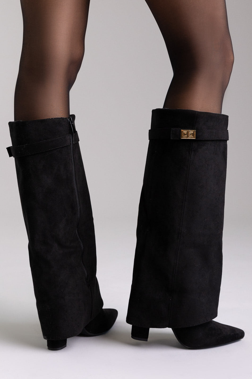 Black suede fold-over knee-high boots with gold buckle and pointed toe, front view on a model in sheer tights