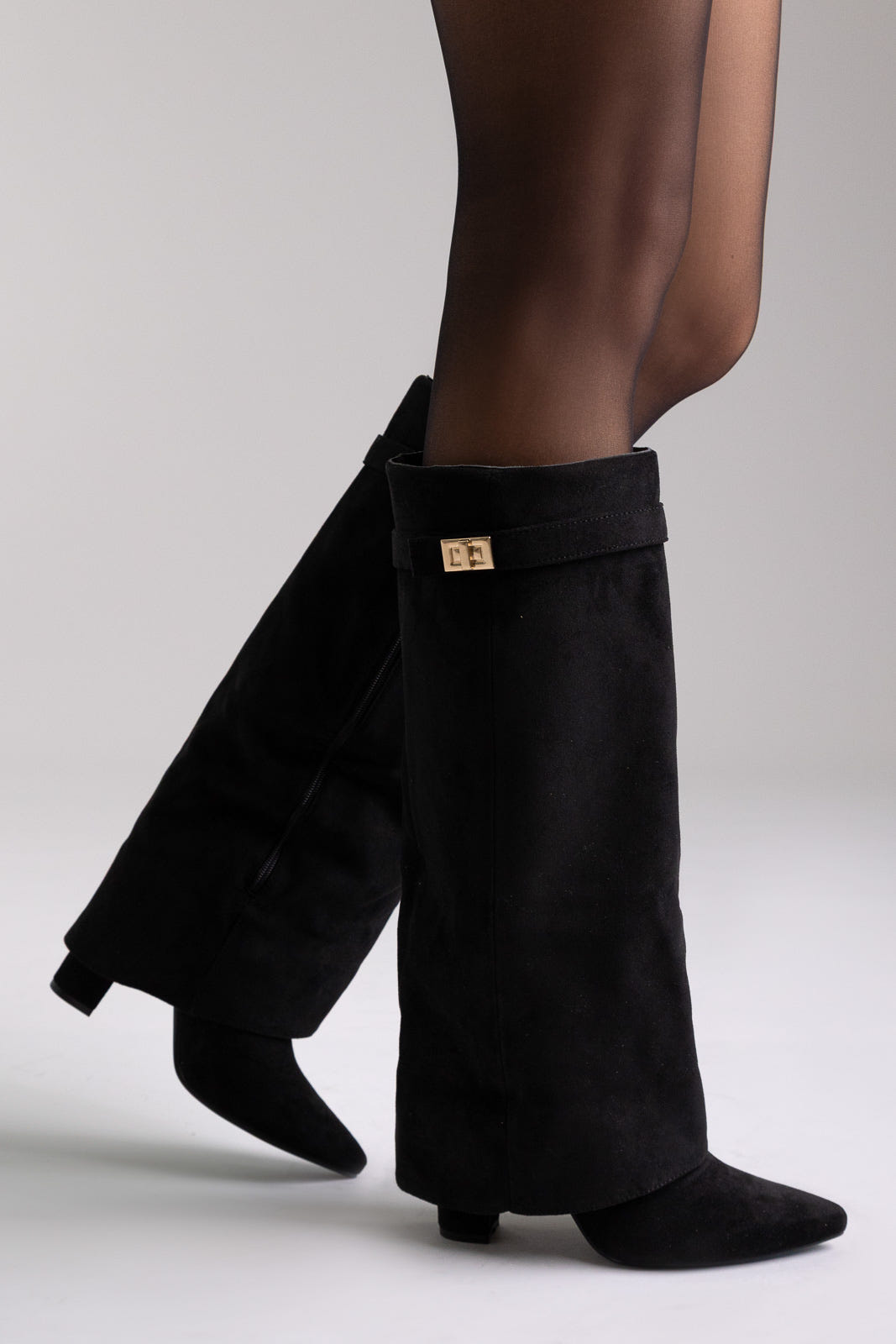 Black suede fold-over knee-high boots with gold buckle and pointed toe, angled view on a model in sheer tights