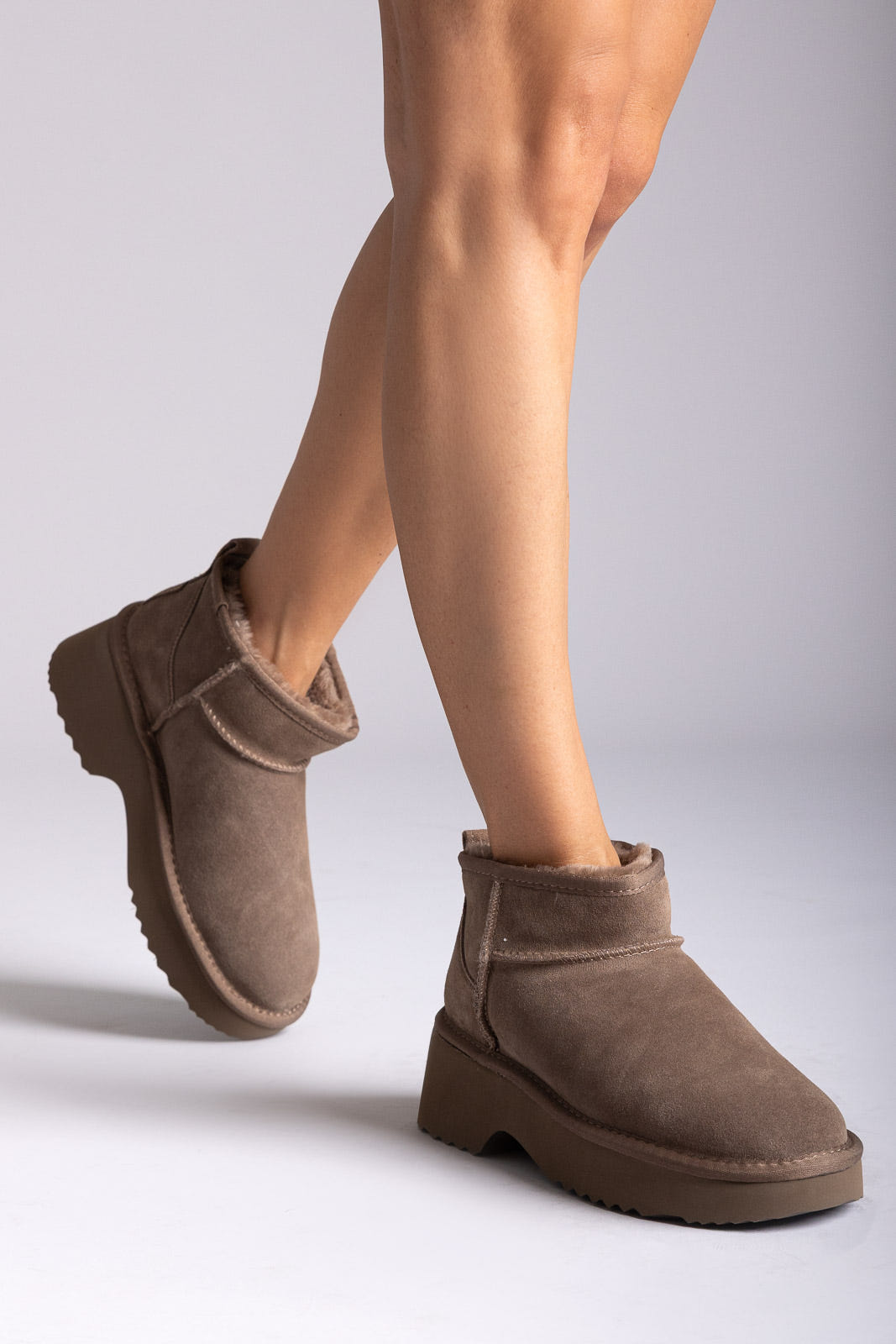 Taupe suede mini boots with fleece cuff and chunky sole, three-quarter view on a model's legs in studio light