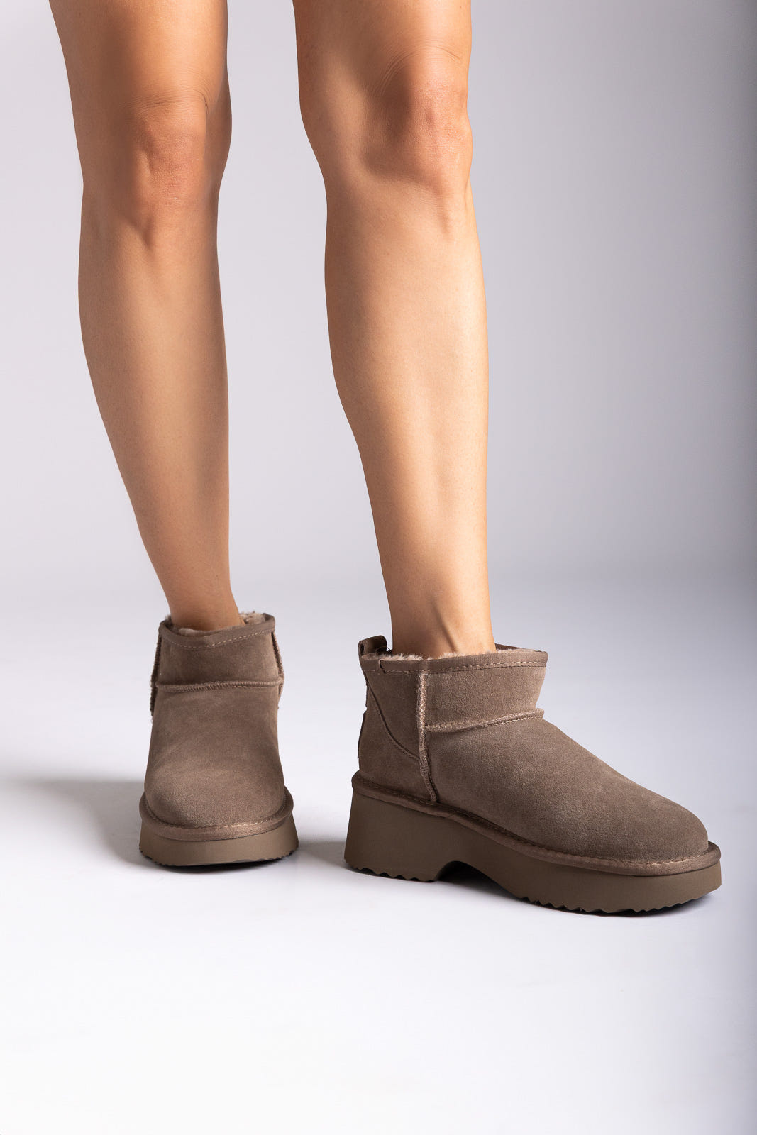 Taupe suede mini boots with fleece lining and chunky rubber sole, front view on a model's legs in studio light