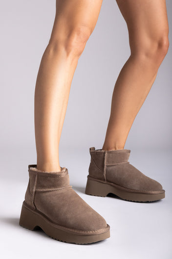 Taupe suede mini boots with fleece lining and chunky sole, side view on a model's legs in studio light