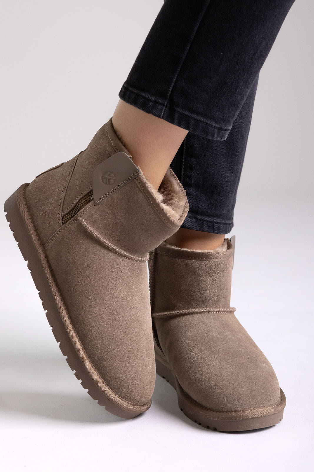 Taupe suede shearling-lined mini ankle boots with pull tab, angled view on a model in jeans