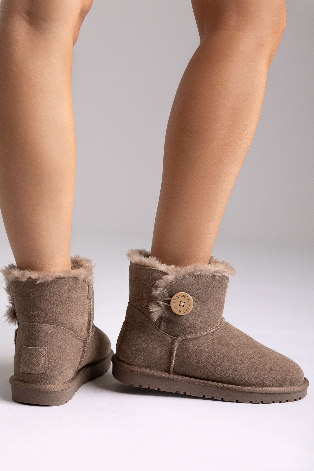 Taupe suede shearling-lined mini ankle boots with button detail, angled view on a model in studio light