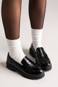 Black patent leather chunky lug-sole penny loafers worn with white socks, angled side view on a model