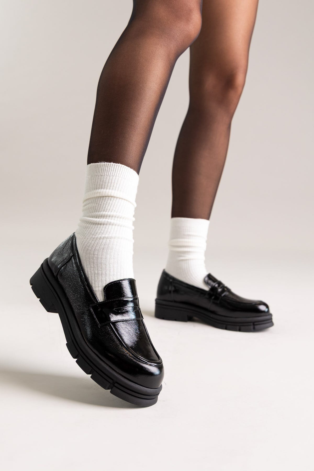 Black patent leather chunky lug-sole penny loafers worn with white socks, side view on a model