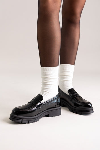 Black patent leather chunky lug-sole penny loafers worn with white socks, angled view on a model