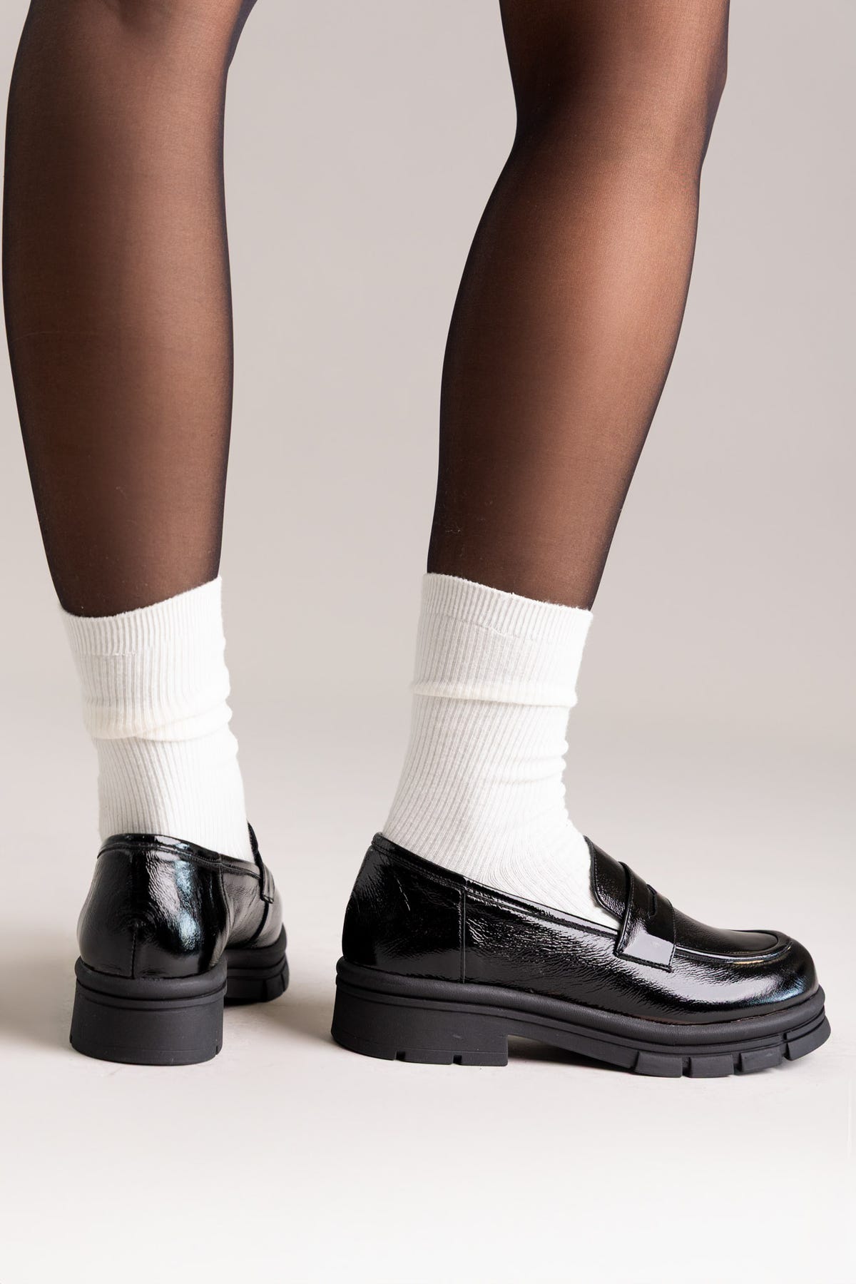 Black patent leather chunky lug-sole penny loafers worn with white socks, rear view on a model