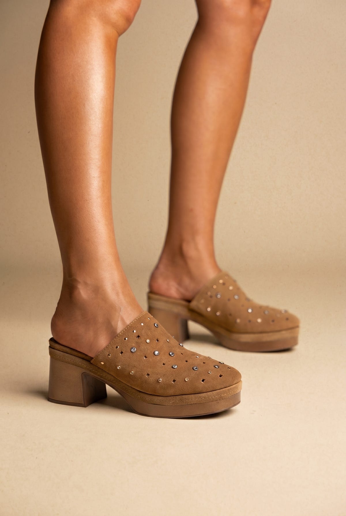 Tan suede clog-style mules with studded uppers and a chunky block heel, worn on a model, three-quarter view