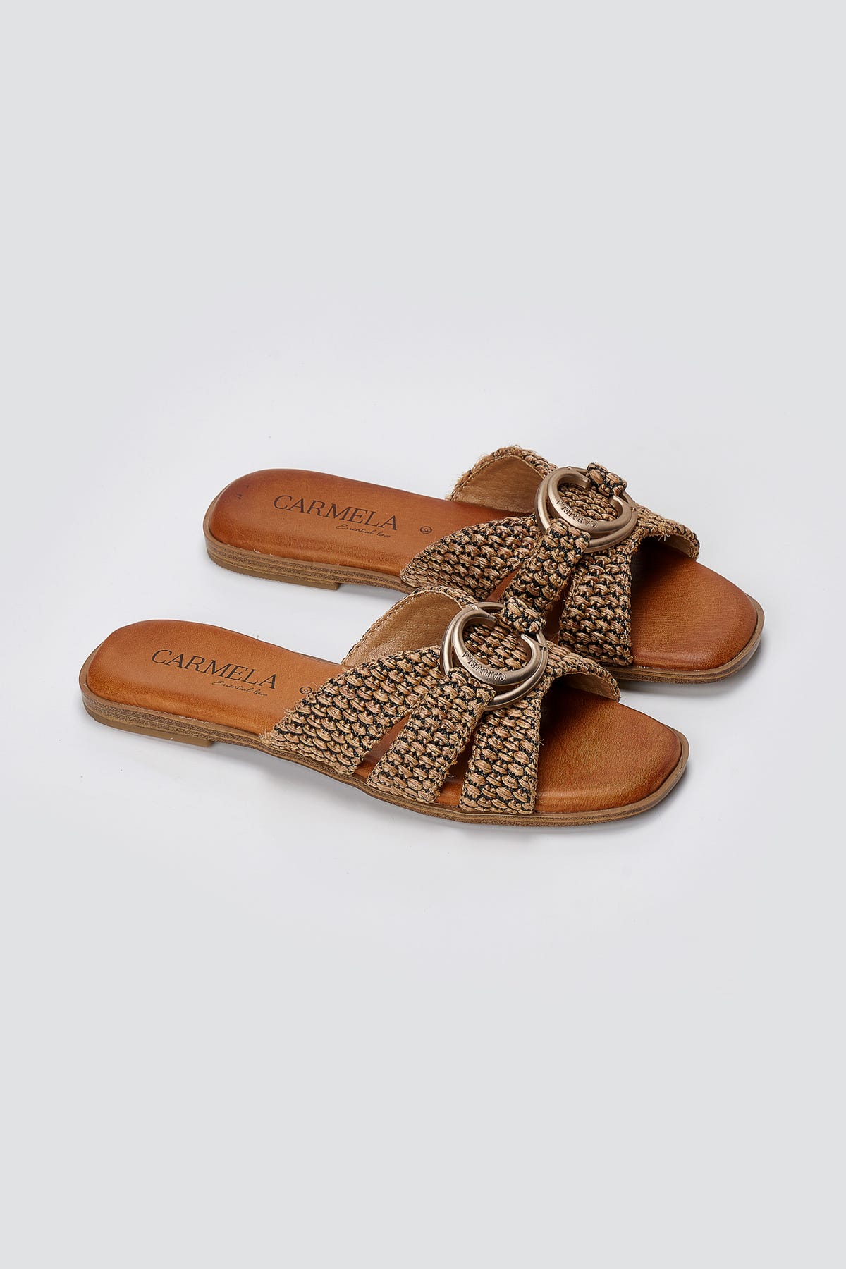 Pair of tan leather flat slides with woven crossover straps and gold ring ornament, three-quarter view
