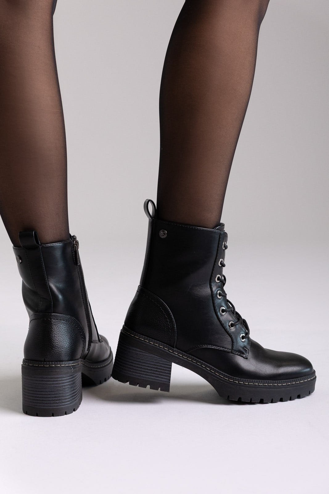 Model legs in black leather lace-up ankle boots with chunky block heels and sheer tights, angled view on a light background
