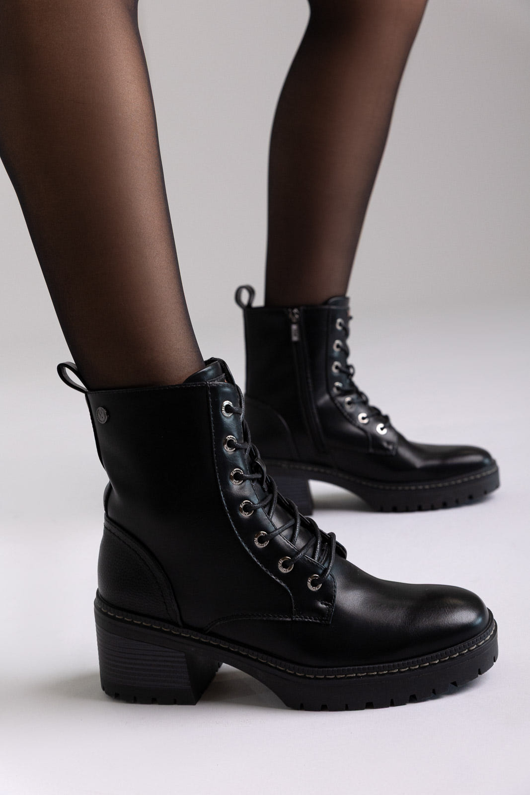 Model legs in black leather lace-up ankle boots with chunky block heels and sheer tights, angled view on a light background