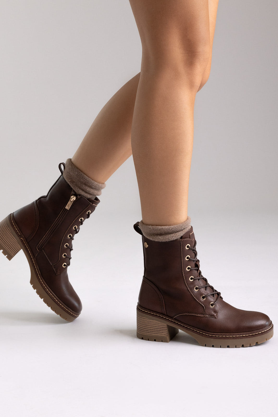 Model legs in brown leather lace-up ankle boots with chunky block heels and cuffed socks, angled view on a light backdrop