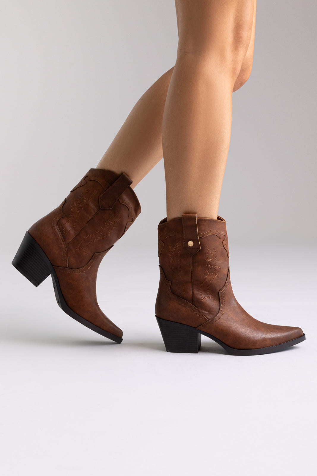 Brown suede western ankle boots with stitched detail and block heel, angled view on a model in studio light