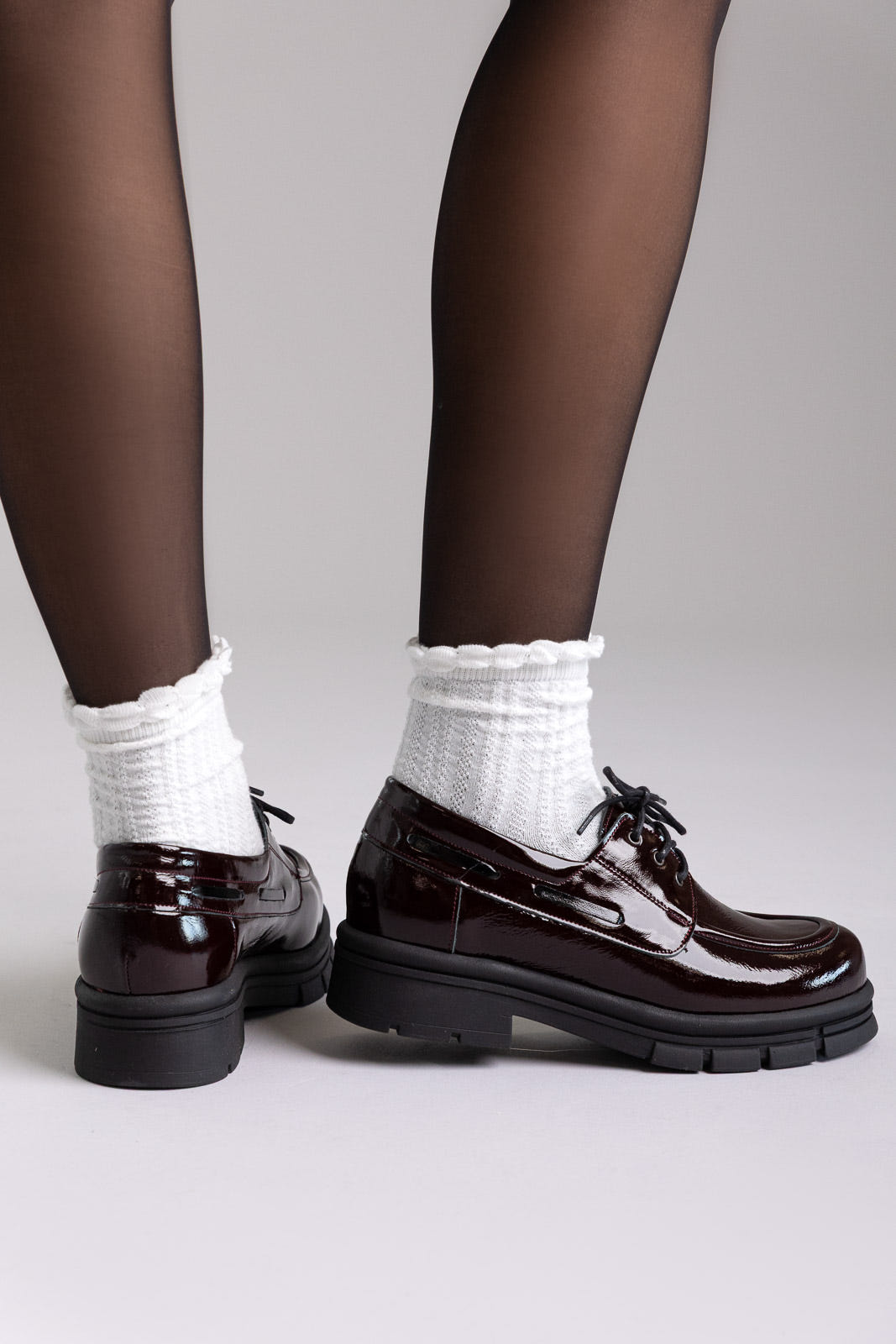 Model legs in burgundy patent chunky sole lace up shoes with white socks, angled view on light grey background