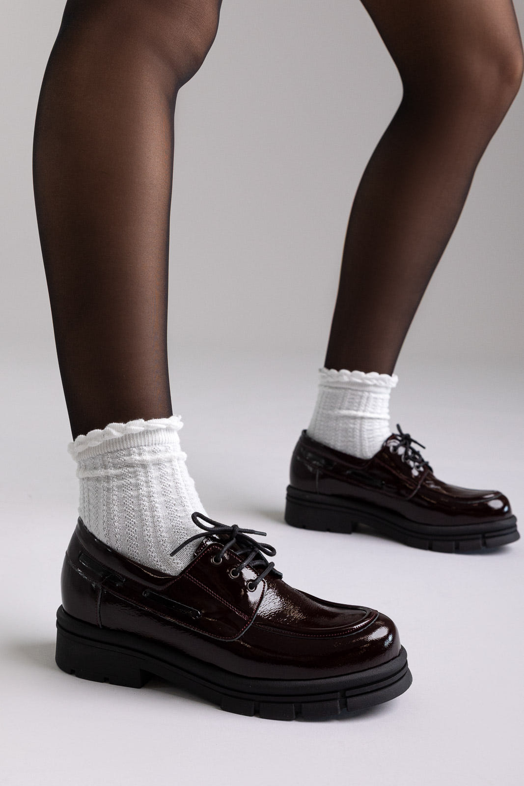 Model legs in burgundy patent chunky sole lace up shoes with white socks, front view on light grey background