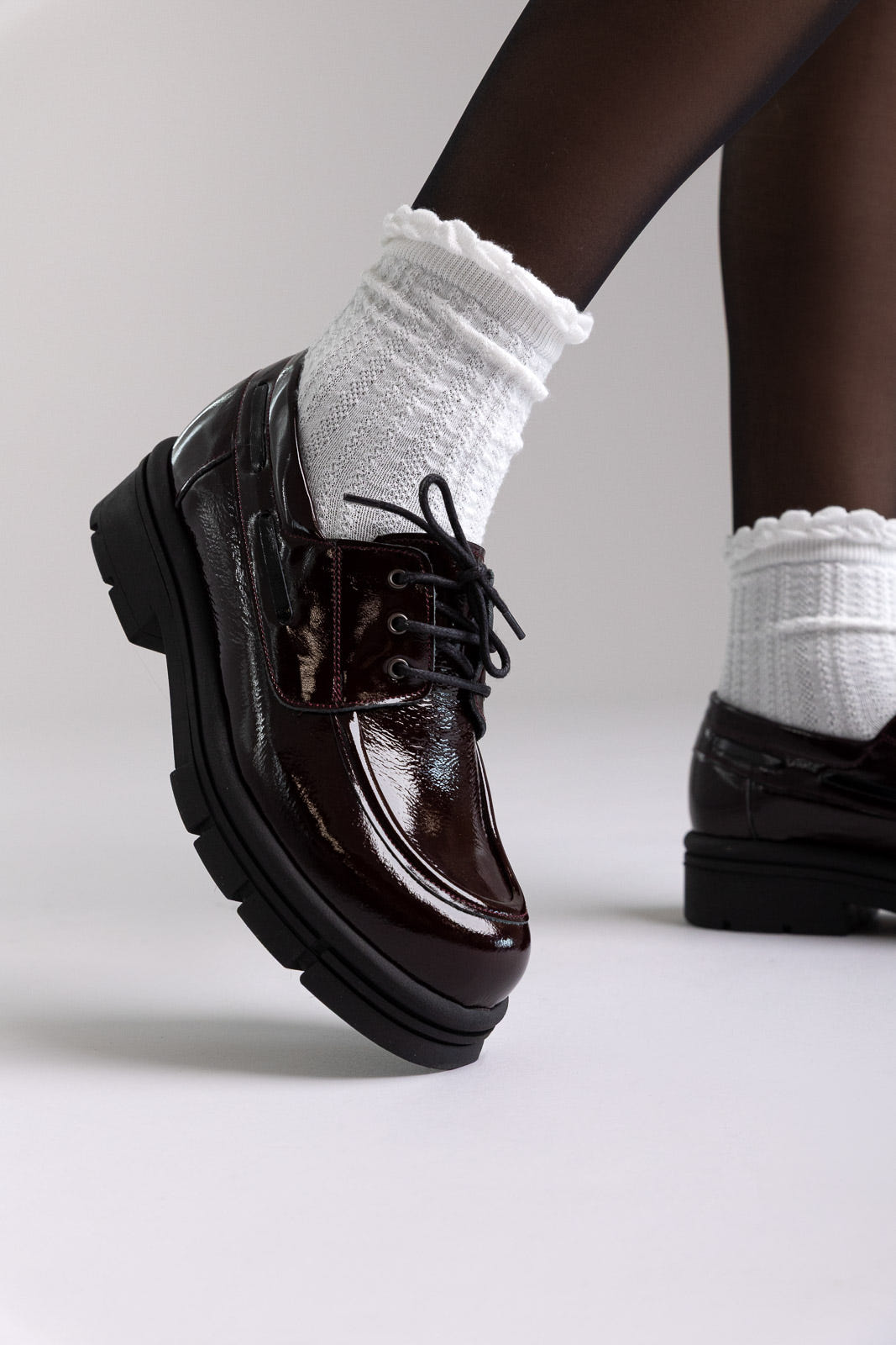 Model legs in burgundy patent chunky sole lace up shoes with white socks, angled view on light grey background