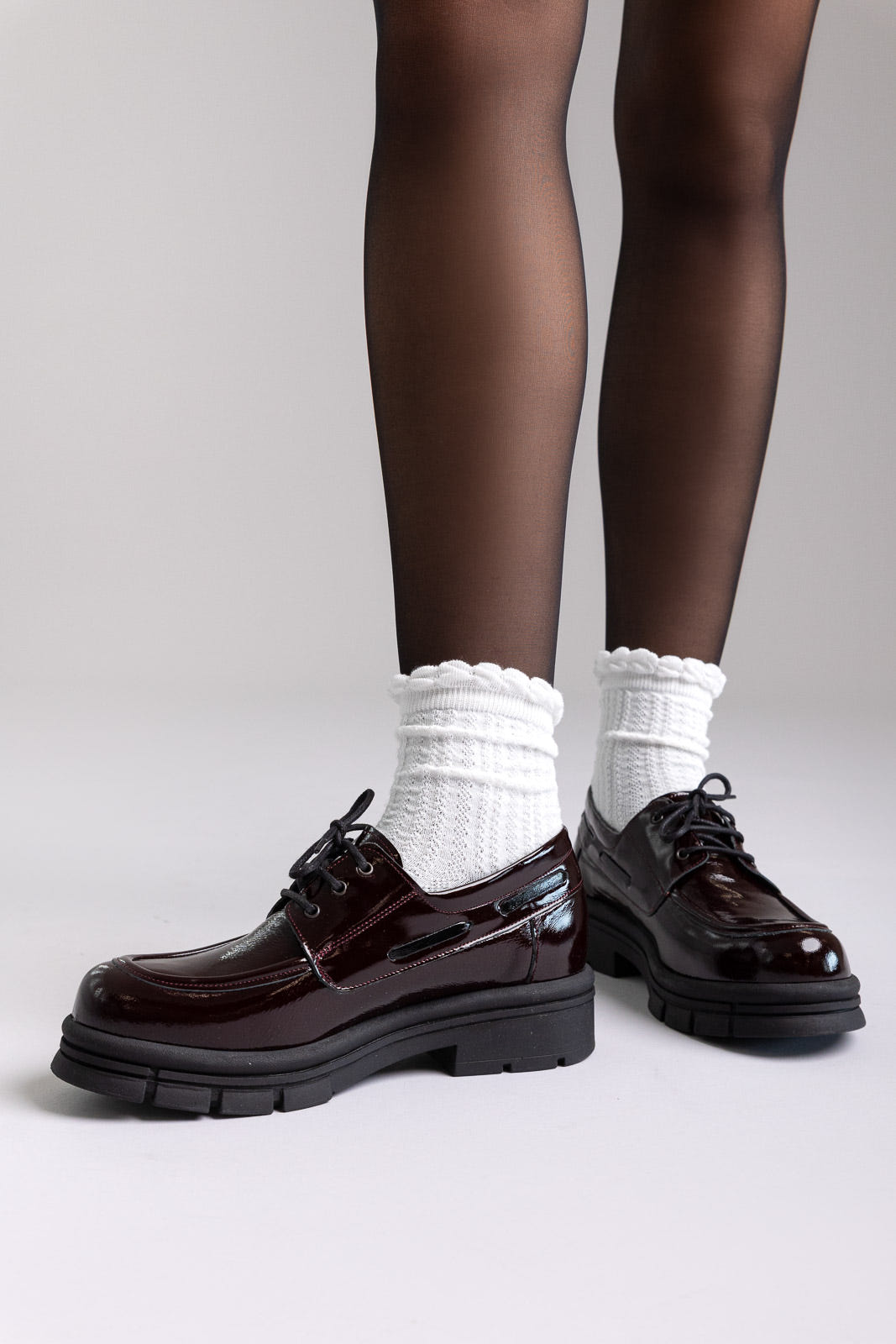Model legs in black patent chunky lace-up shoes with white socks, front angled view on a light grey background