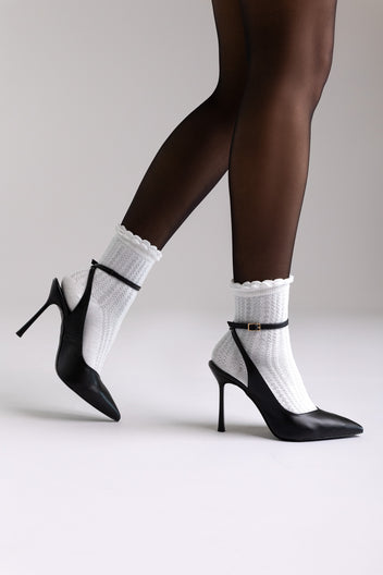 Black leather ankle-strap stiletto pumps with pointed toe, side view on a model with white ruffle socks