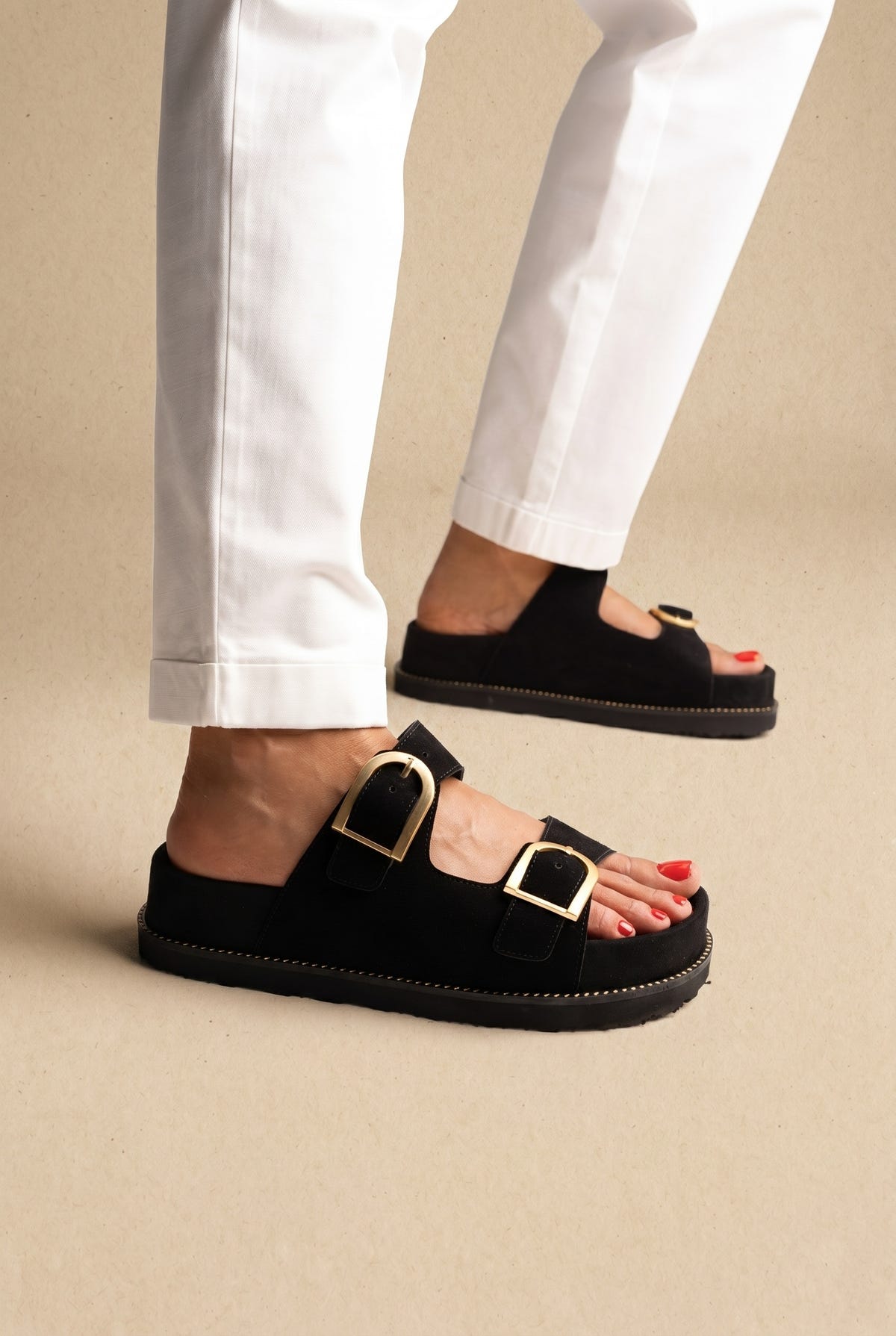 Black suede double-strap platform slides with large gold buckles on a model with white trousers, side view