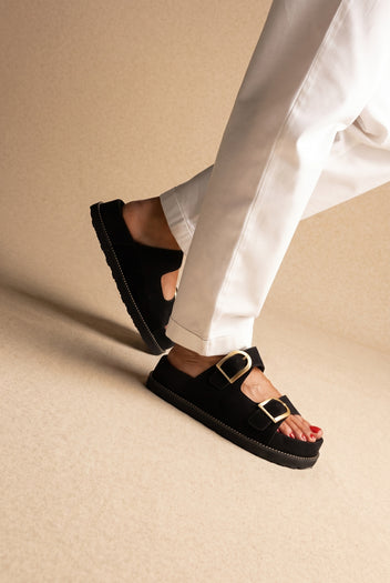 Black suede platform slide sandals with two gold buckles, worn on a model with white trousers, angled view
