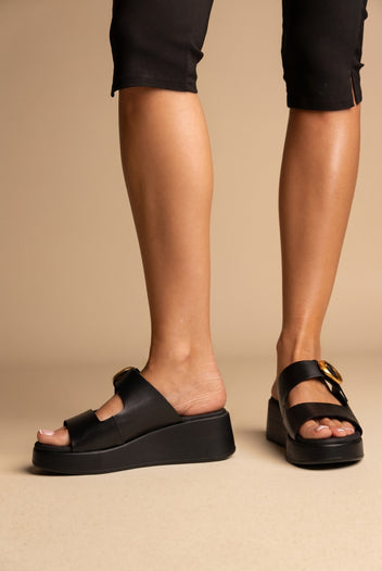 Black leather wedge platform slides with a gold round buckle on a double strap, worn on a model, front view