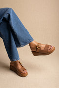 Tan smooth leather platform wedge slide sandals with a wide strap and gold buckle, worn on a model with blue jeans