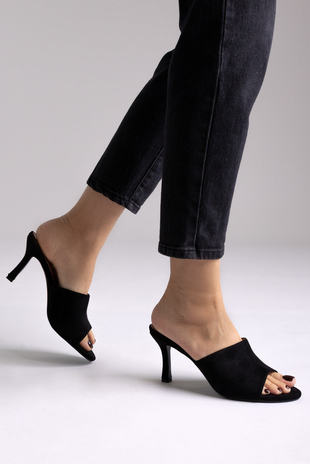 Black suede peep-toe kitten-heel mules, angled view on a model in black jeans in studio light